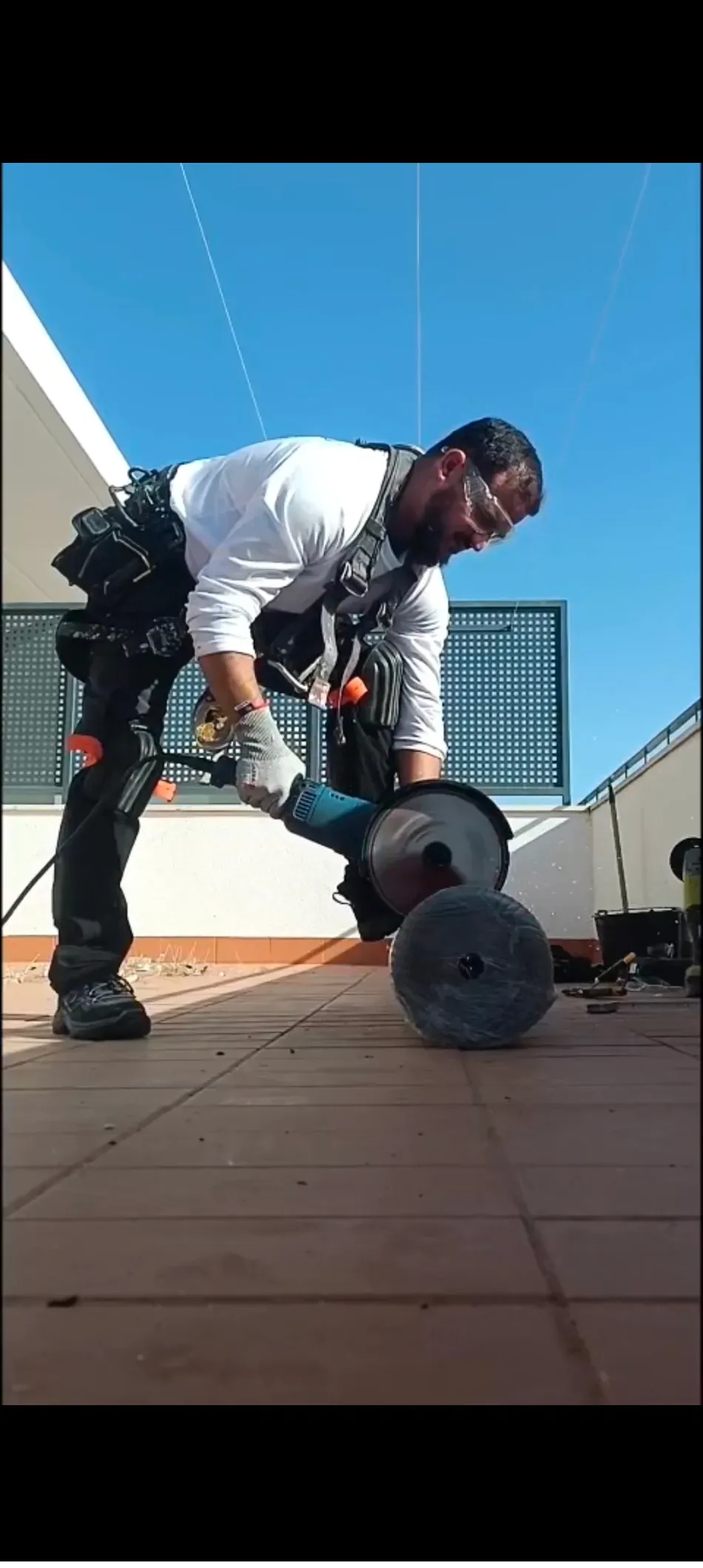 Una persona con ropa de trabajo utiliza una herramienta eléctrica al aire libre sobre una superficie pavimentada bajo un cielo azul.