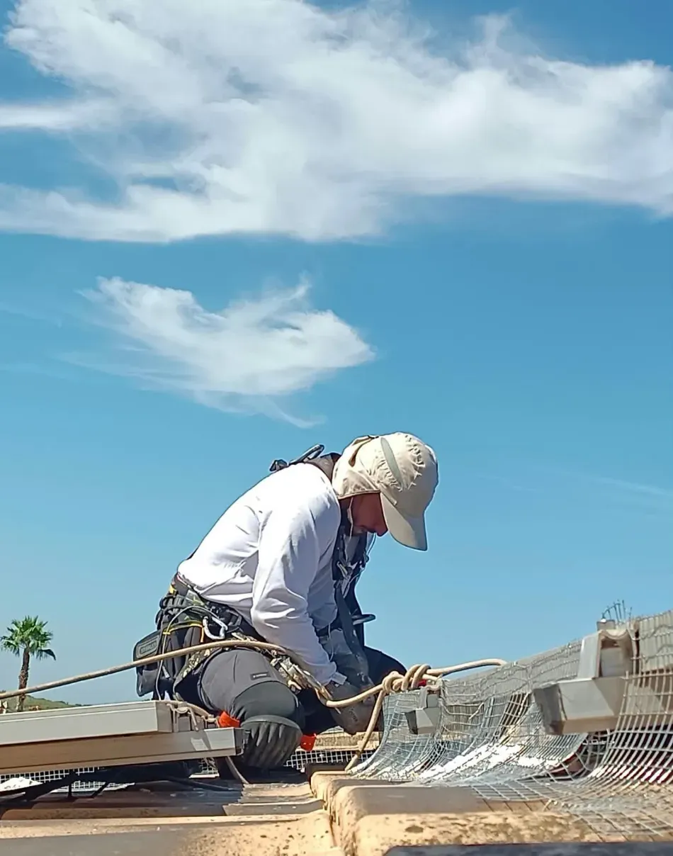 Un trabajador con gorro blanco se arrodilla en una azotea, manipulando una red bajo un cielo azul con nubes.