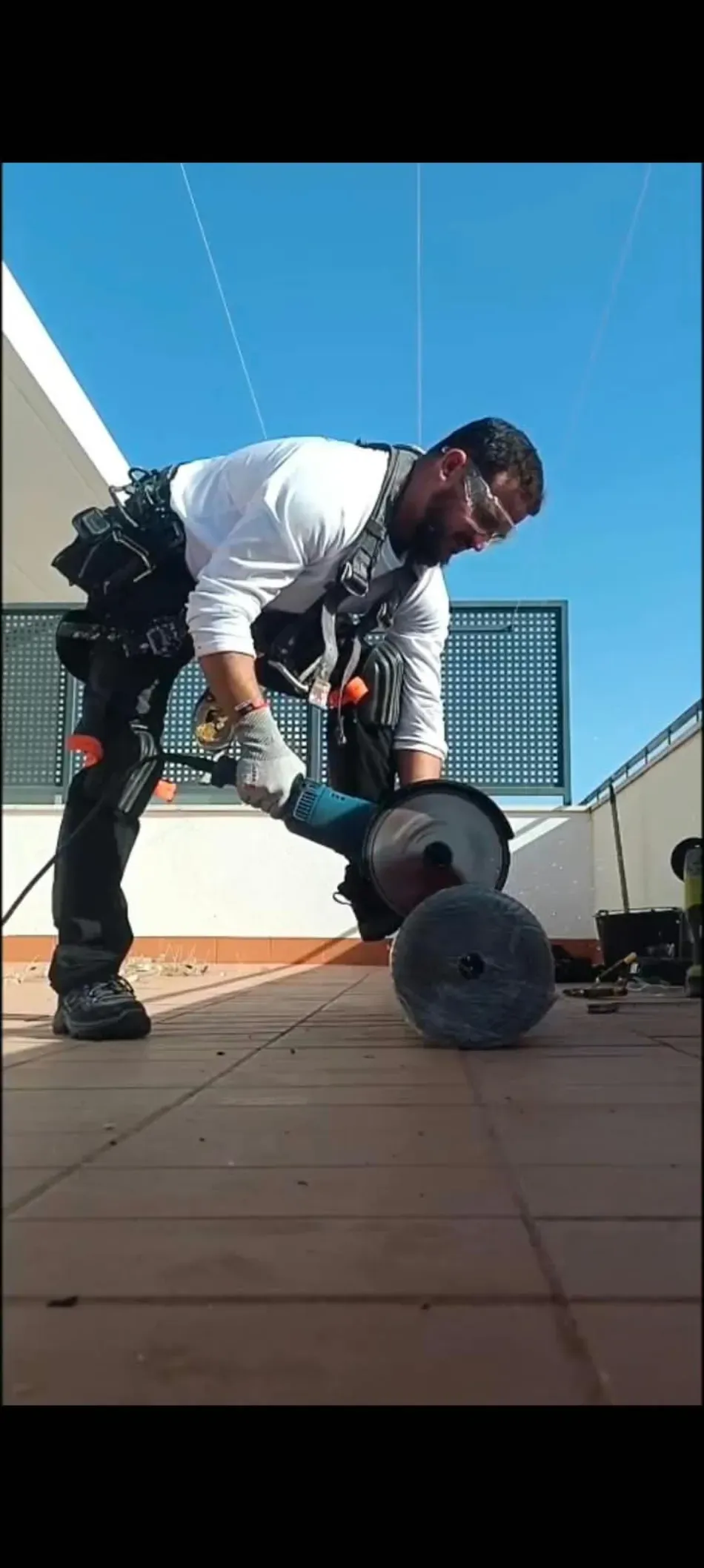 Hombre con ropa de trabajo inclinado para ajustar una máquina con ruedas en una azotea bajo un cielo azul despejado.