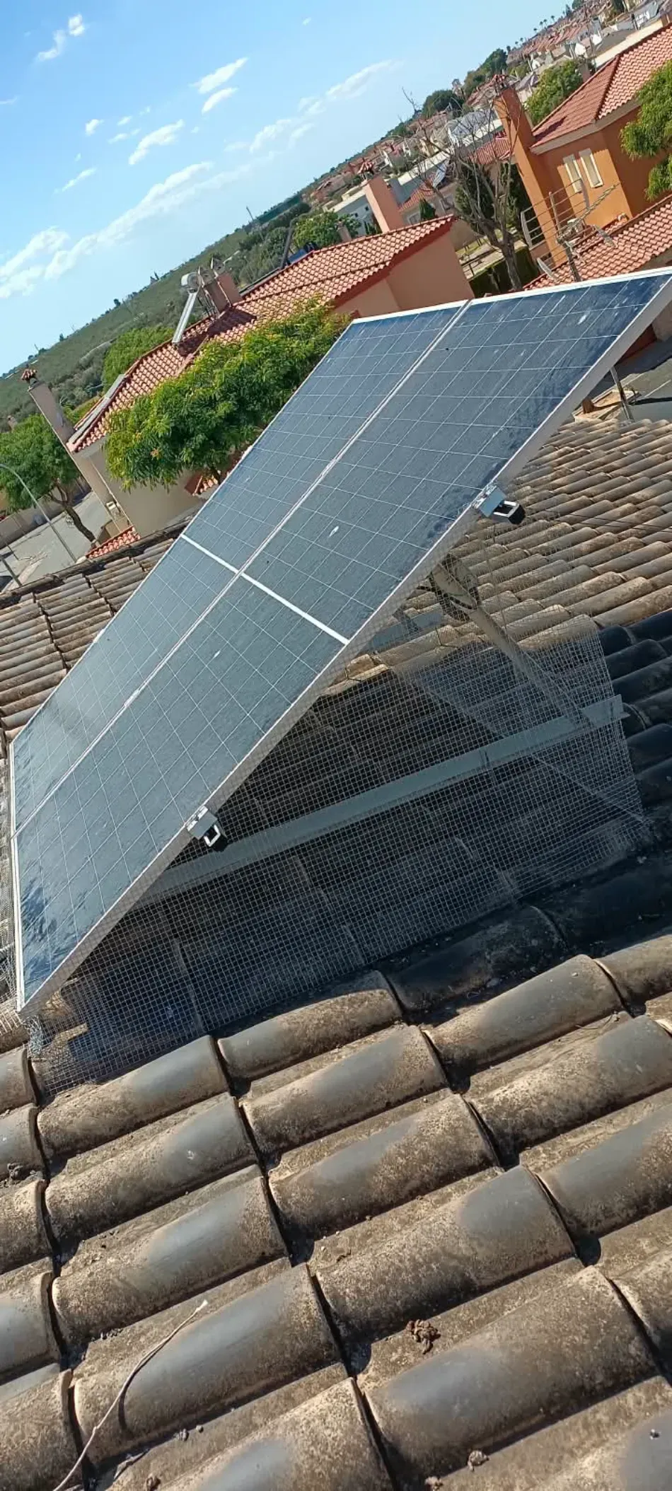 Panel solar montado en un tejado sobre tejas de arcilla roja, con casas del vecindario al fondo.