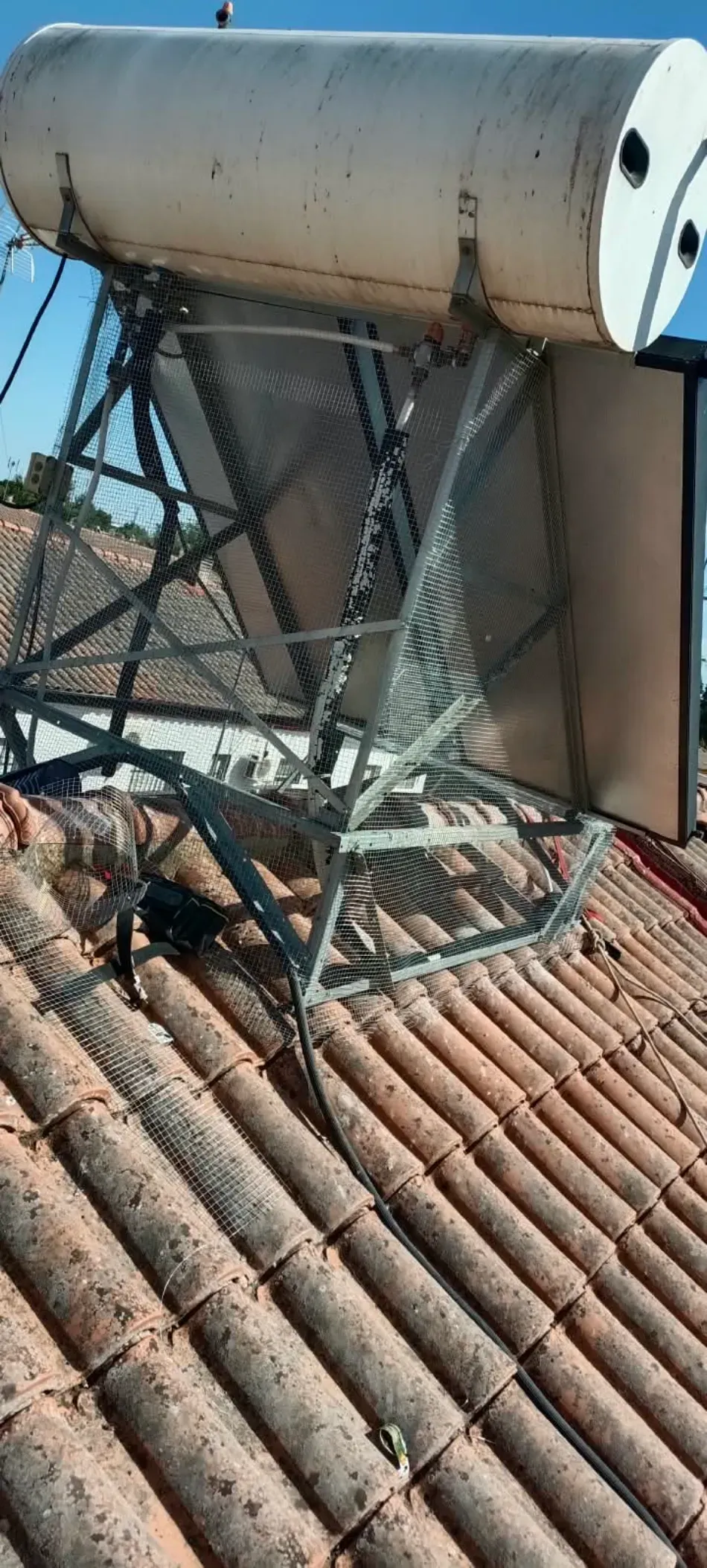 Tanque de calentador de agua solar y estructura de soporte sobre un tejado de tejas rojas bajo un cielo azul.