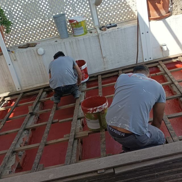 Dos trabajadores pintan una superficie roja sobre una estructura de madera, utilizando cubos, al aire libre.