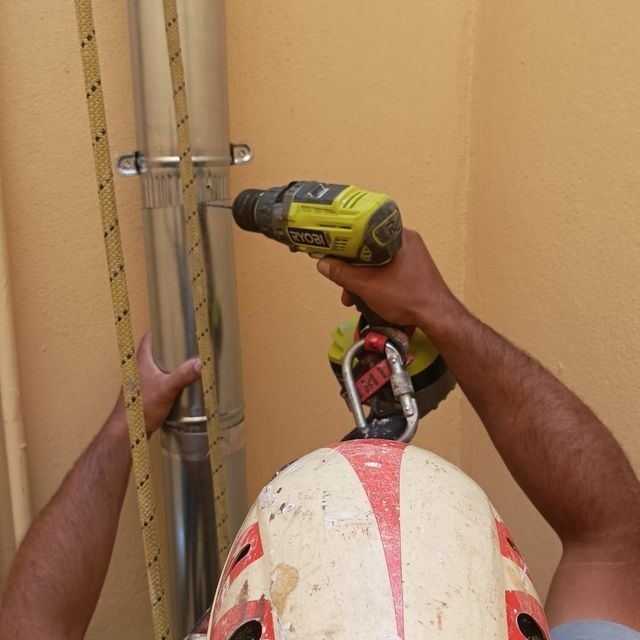 Persona con casco utilizando un taladro en una tubería metálica, sujeta con una cuerda. Fondo de pared amarilla.