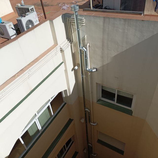 Tuberías metálicas verticales en el exterior de un edificio, que descienden pasando por ventanas y una pared de color amarillo pálido.