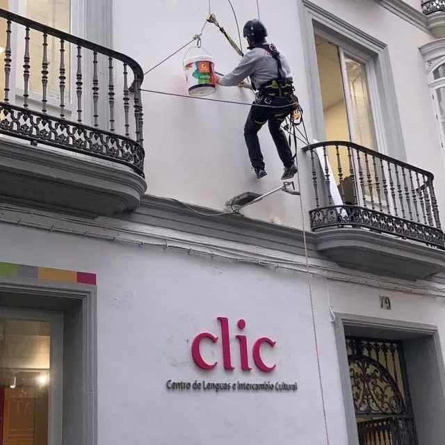 Una persona pinta la fachada de un edificio mientras está suspendida por cuerdas. El edificio dice 