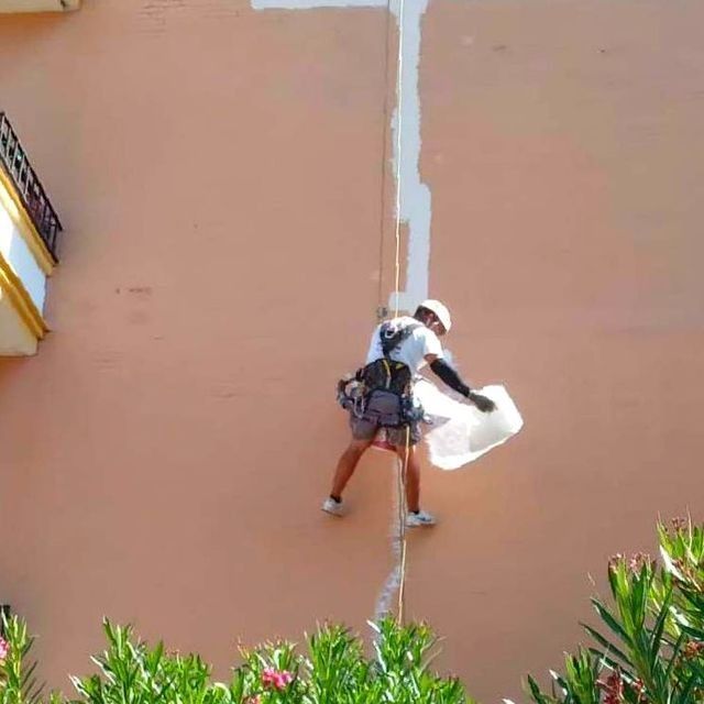 Hombre suspendido de una cuerda, aplicando sellador blanco a la pared exterior del edificio.