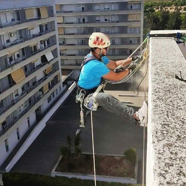 Un trabajador que realiza rápel desciende de un edificio. Lleva casco, arnés y camisa azul, sujetos con cuerdas.
