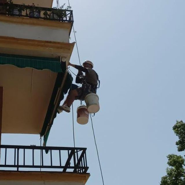 Persona sujeta con cuerdas, con arnés y cubos de pintura, pinta la fachada de un edificio. Cielo despejado.