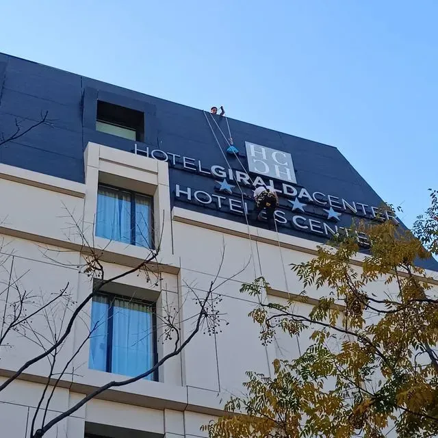 Obreros suspendidos de cuerdas instalan letras en el letrero del Hotel Giralda Center, un edificio alto, contra un cielo azul.