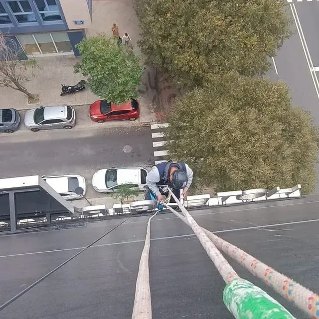 Persona descendiendo en rappel por un edificio, sujetando cuerdas. Calle y coches aparcados abajo.