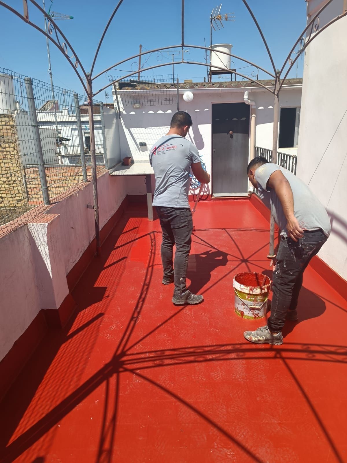 Dos personas pintan una terraza roja en la azotea. Una pinta, la otra observa, bajo un arco metálico. Sol radiante.