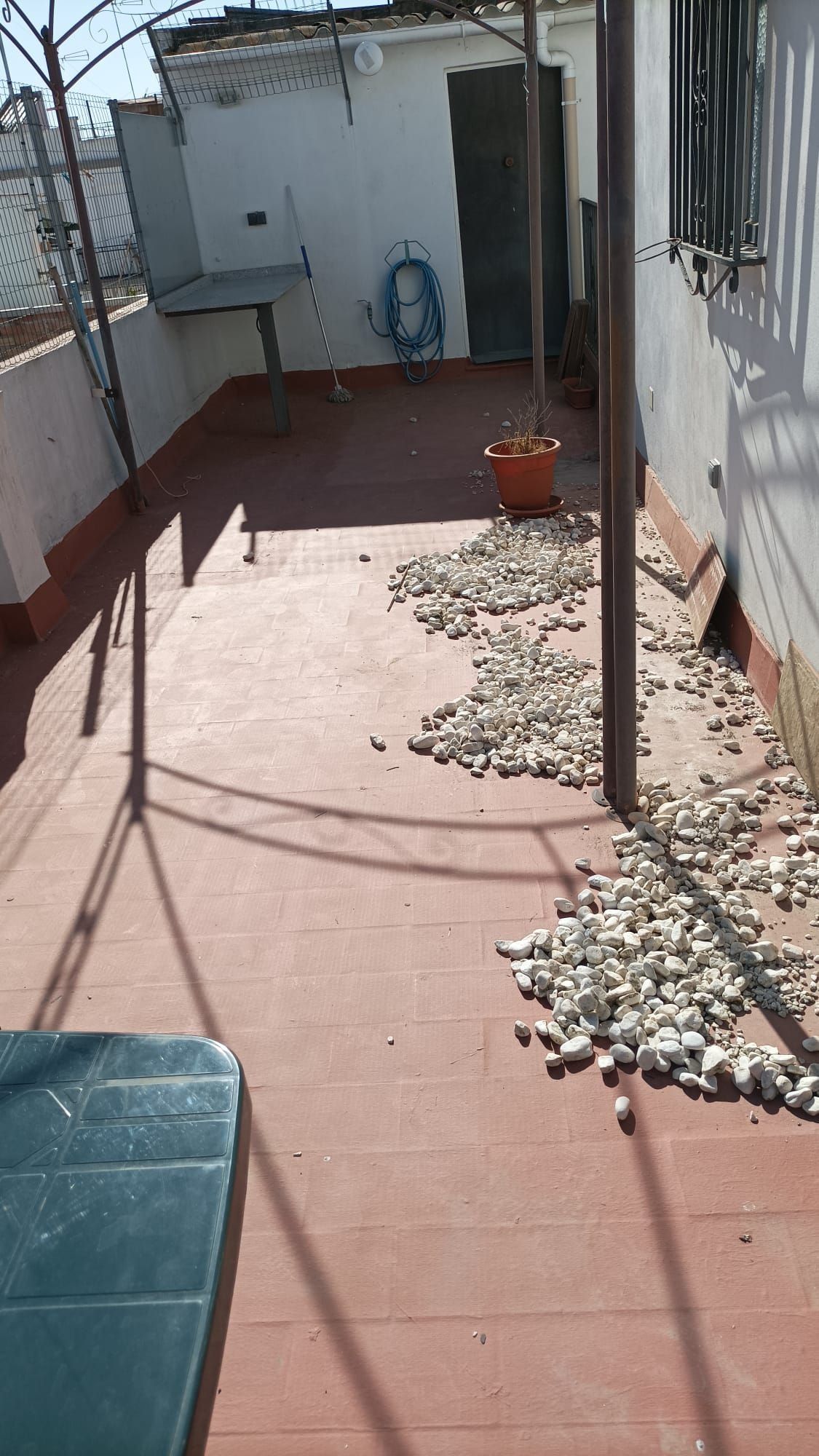 Terraza en la azotea con suelo rojo, rocas grises y algunas plantas en macetas; sombras de la estructura superior.