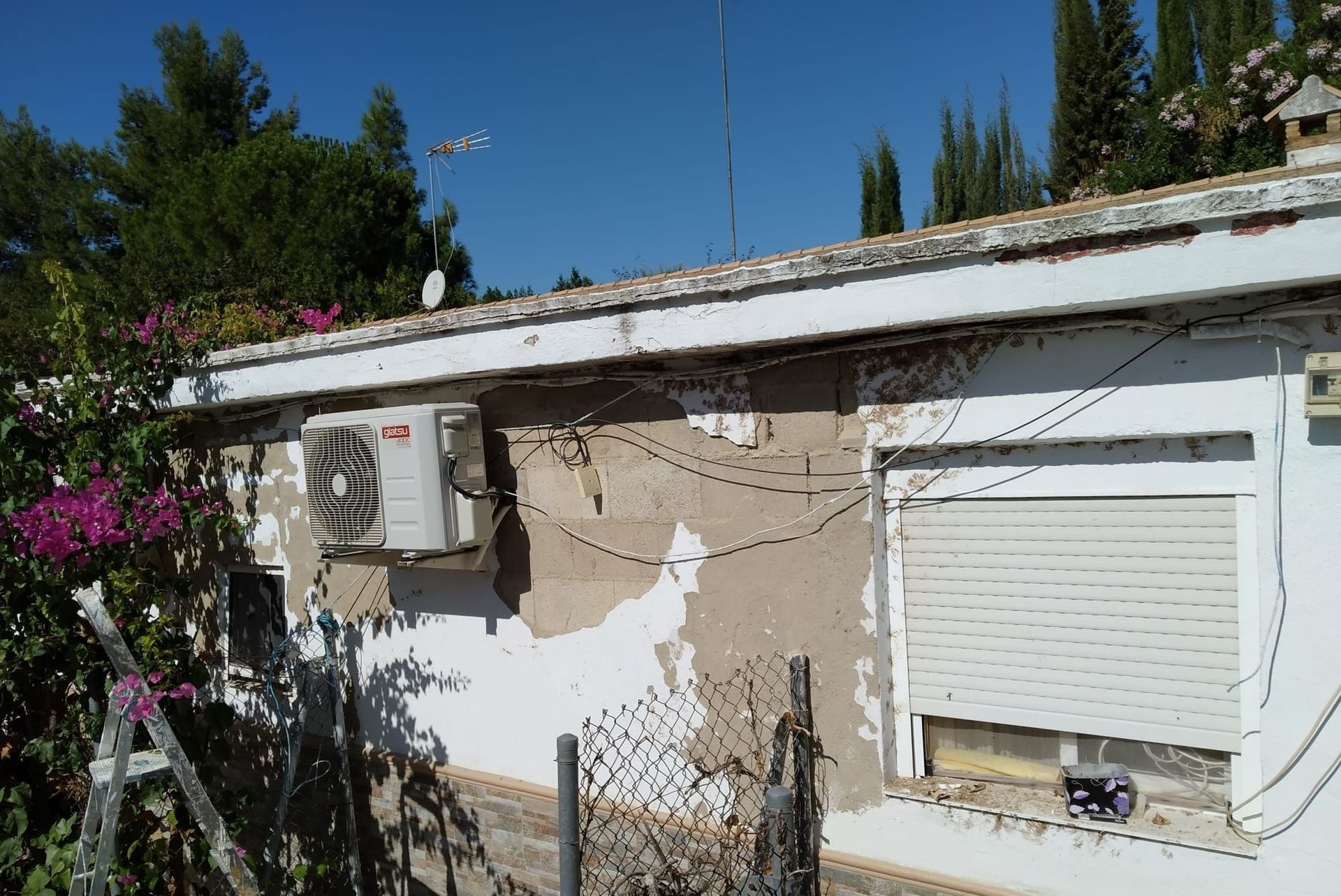 Vista exterior de un edificio con pintura desconchada, una unidad de aire acondicionado y una ventana cerrada con persiana enrollable.
