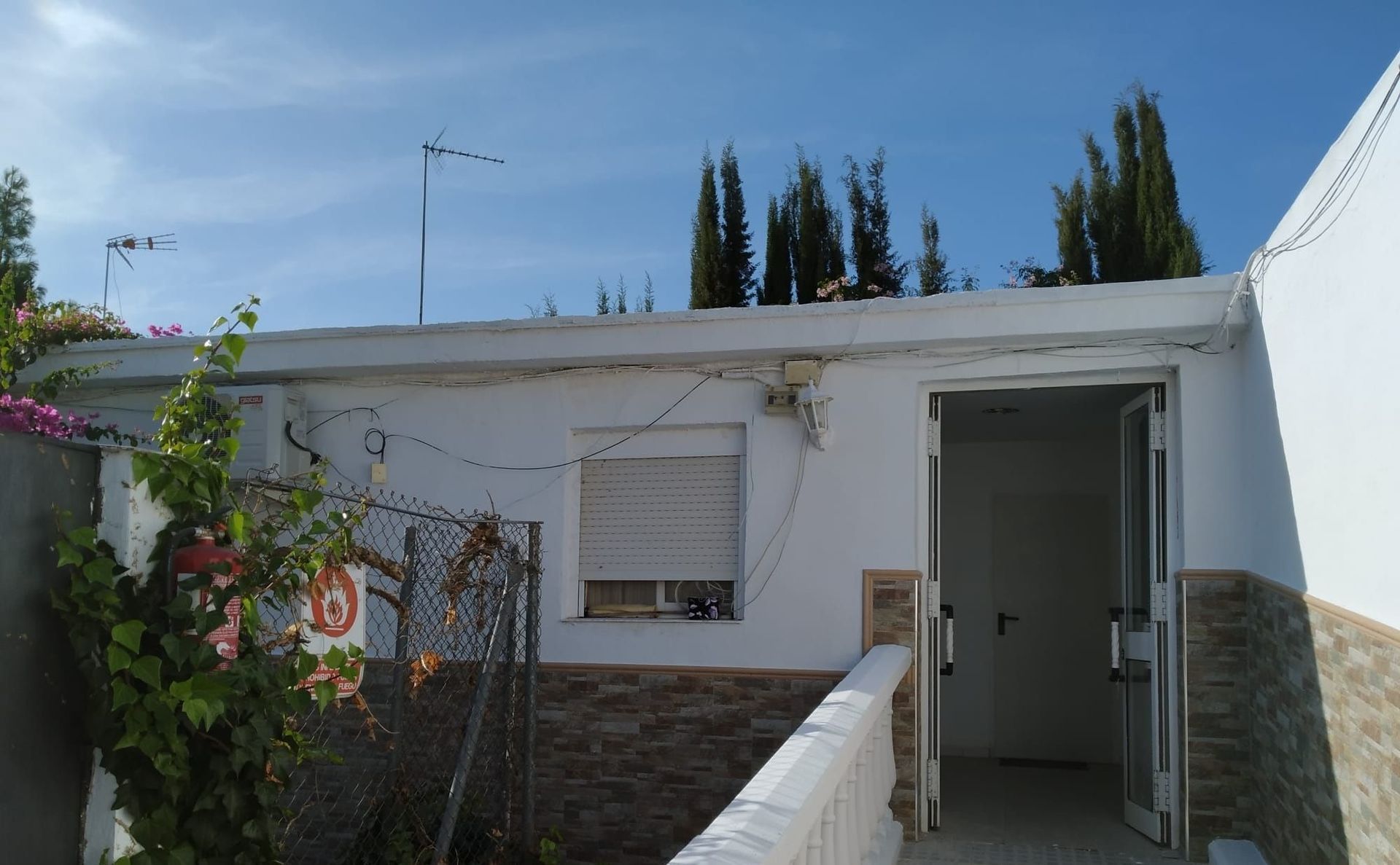 Edificio blanco con puerta abierta, ventana pequeña y antenas contra un cielo azul.