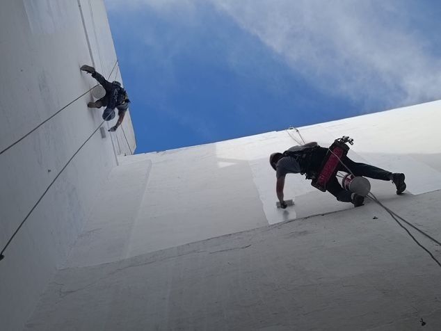 Dos personas sujetas a cuerdas, pintando el exterior de una estructura cilíndrica blanca contra un cielo azul.
