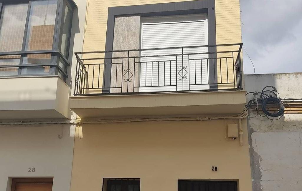 Fachada de un edificio de tres plantas con balcón. Paredes de color amarillo pálido, molduras oscuras y contraventana blanca cerrada.