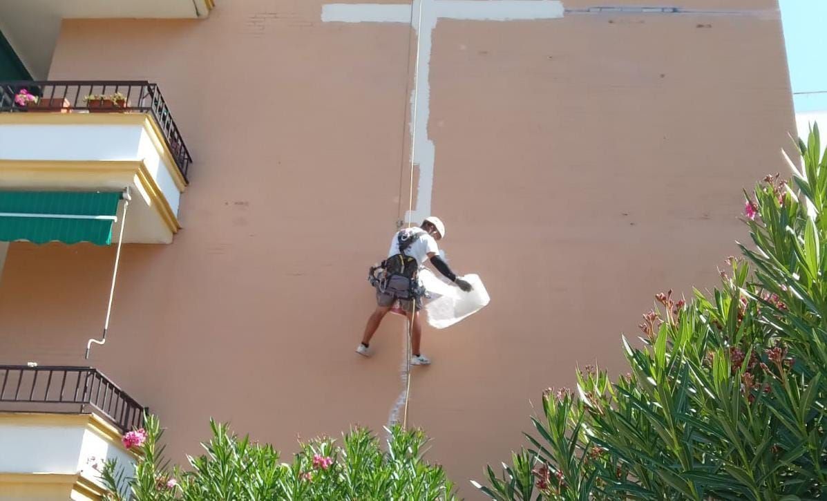 Una persona suspendida por cuerdas, pintando una línea blanca sobre la fachada de un edificio color melocotón.
