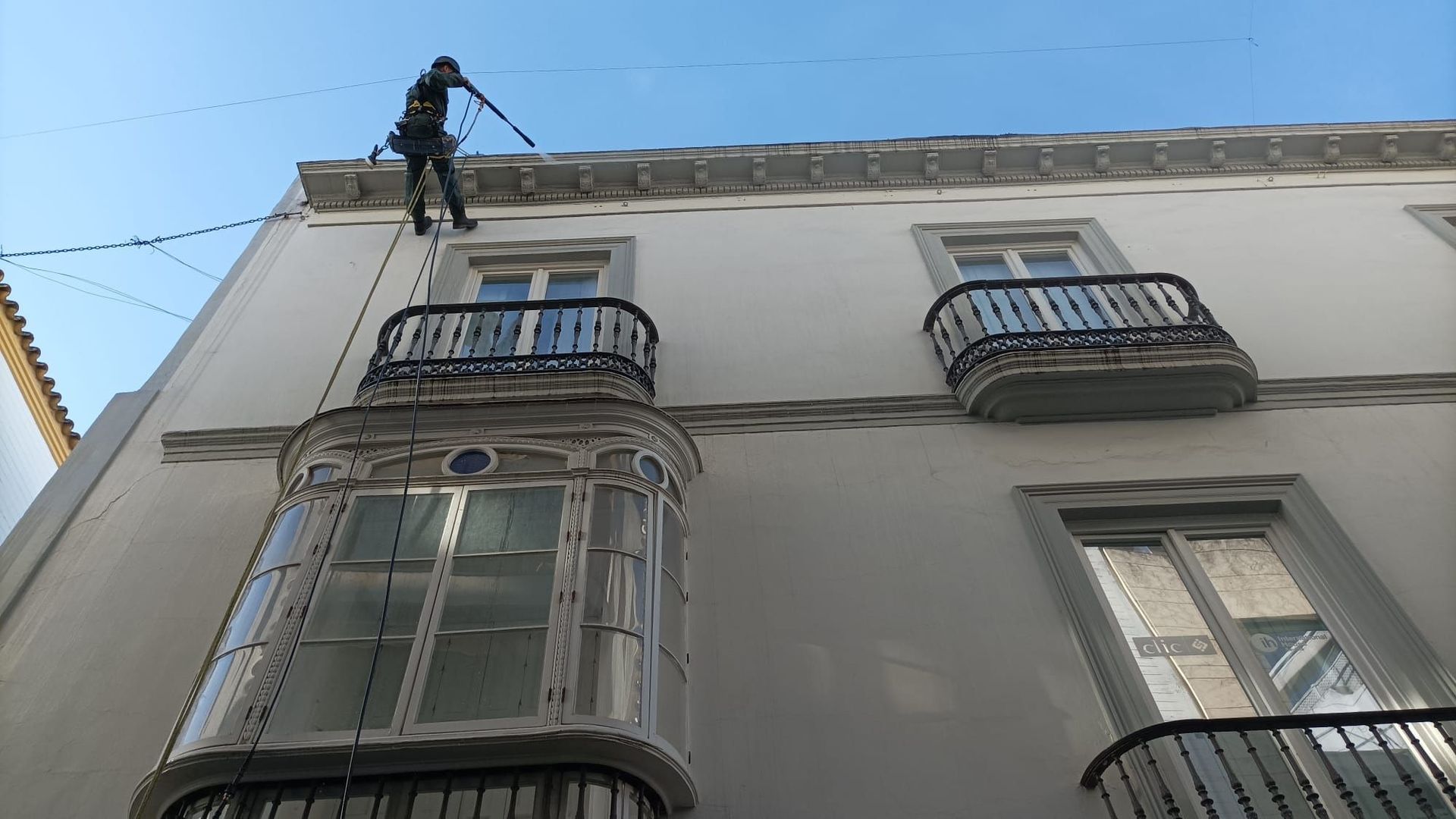 Una persona sujeta a una cuerda repara la fachada del edificio, dos balcones y un mirador en la planta baja.