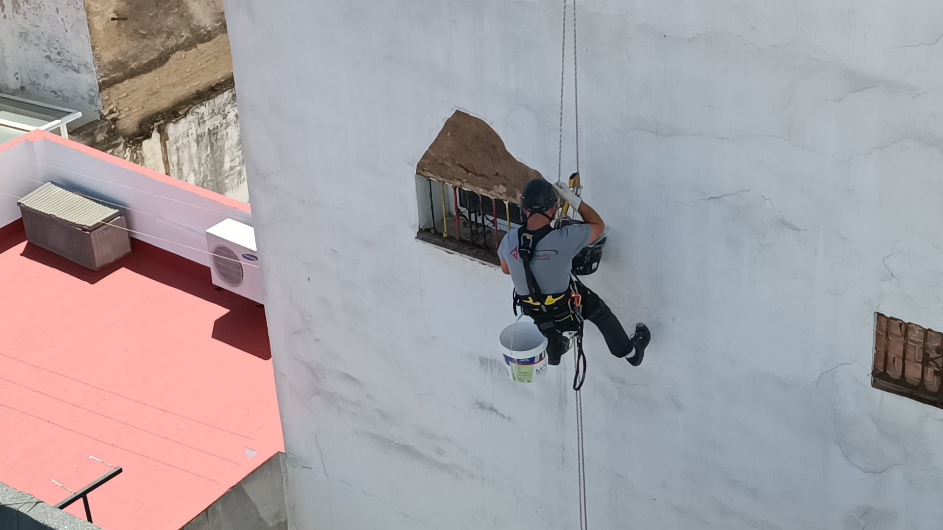 Hombre pintando una pared exterior, suspendido por cuerdas, cerca de una ventana y del tejado.