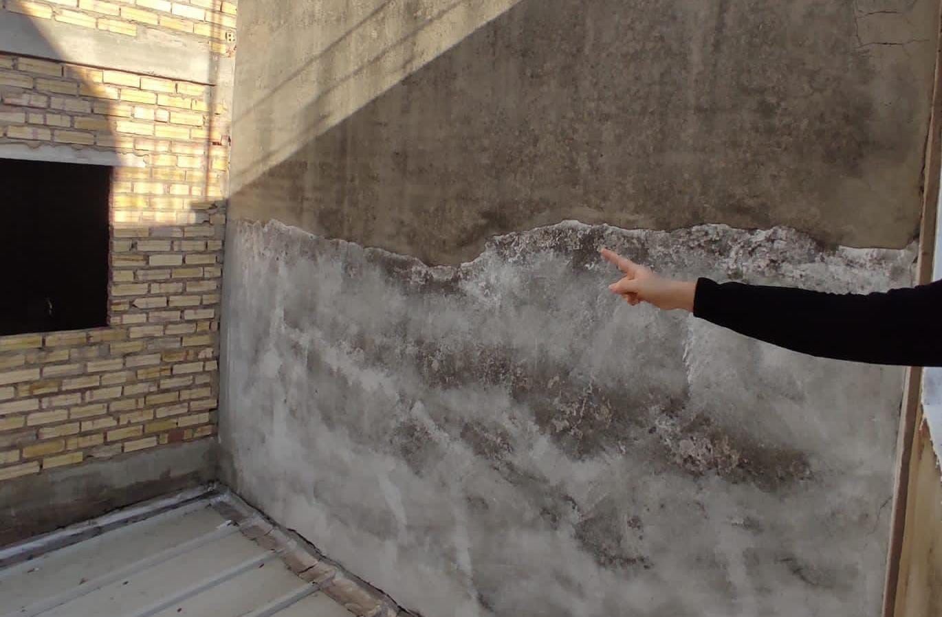 Mano señalando una pared de hormigón húmeda y texturizada. La luz del sol proyecta sombras. Pared de ladrillo adyacente con una ventana negra.