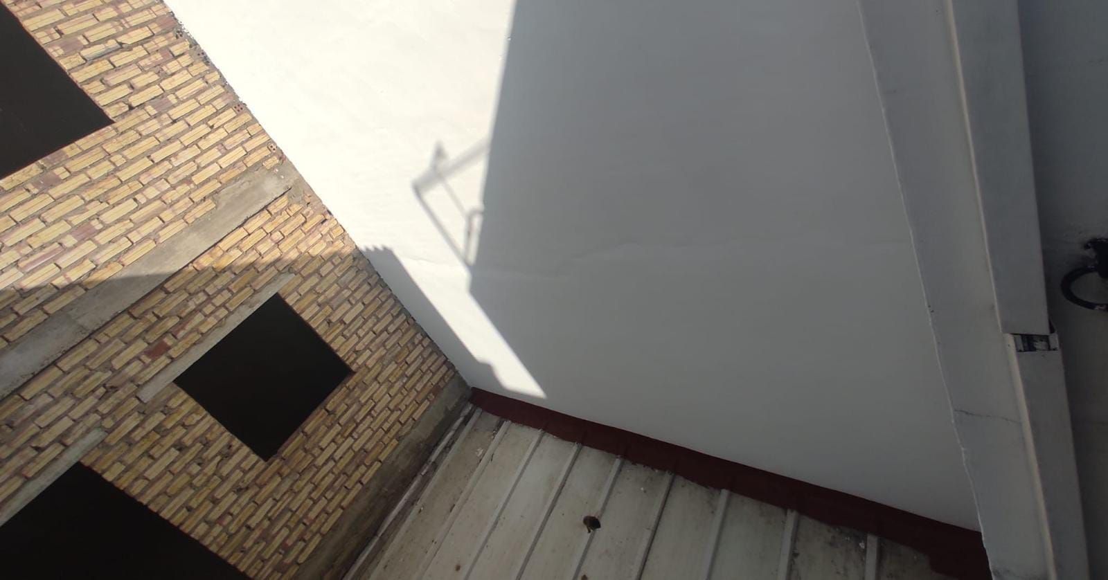 Vista desde arriba de una esquina con pared de ladrillo, pared blanca y techo gris. La luz del sol proyecta sombras.