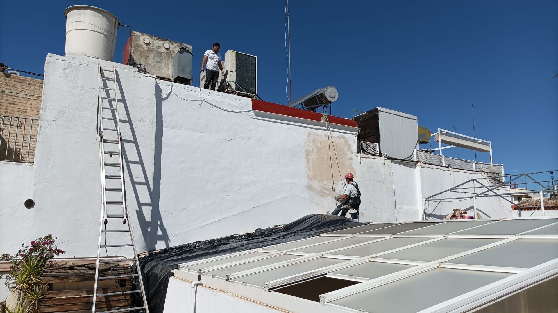 Personas trabajando en una azotea con equipo y una escalera, apoyadas contra una pared blanca bajo un cielo azul.