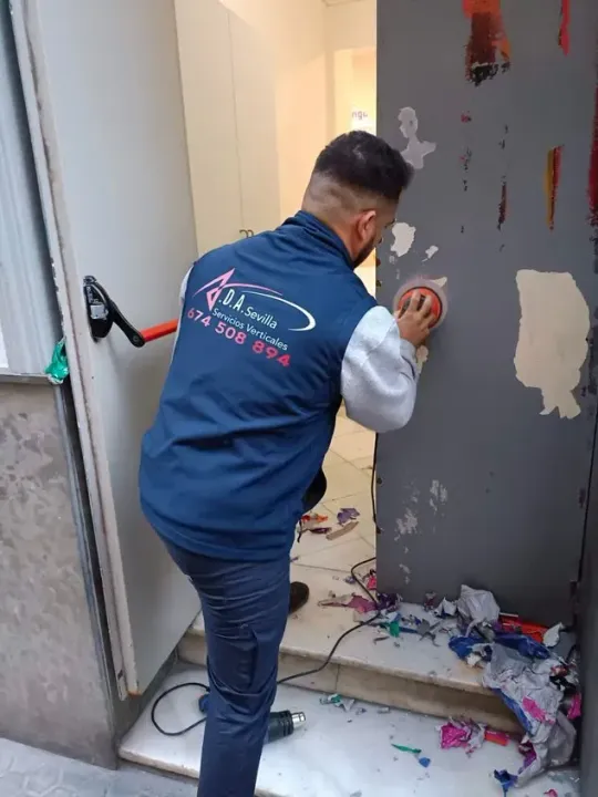 Hombre lijando una puerta, con chaleco azul, de pie en un umbral con escombros en el suelo.