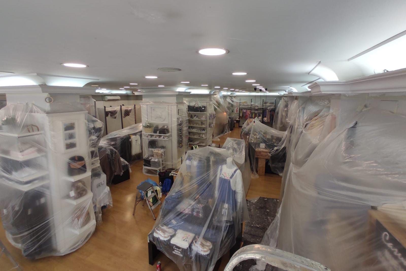 Interior de una tienda; artículos y muebles cubiertos con plástico para su protección. Filas de estanterías y expositores.