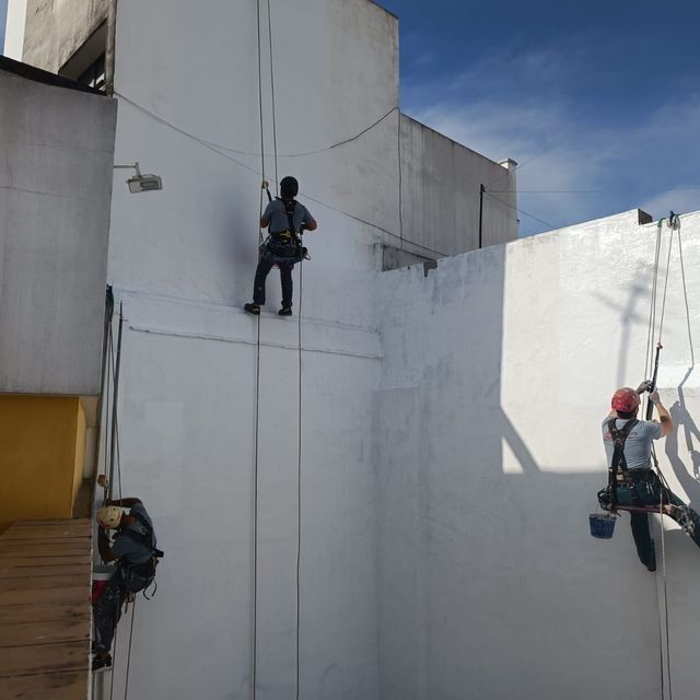 Tres trabajadores sujetos con cuerdas, pintando el exterior de un edificio blanco en un día soleado.
