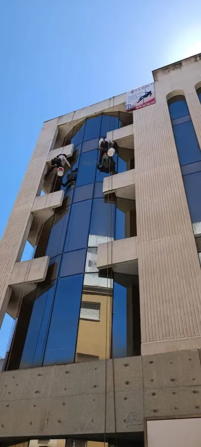 Dos limpiadores de ventanas sujetos con cuerdas limpian un edificio de varias plantas con ventanas de cristal reflectante bajo un cielo azul.