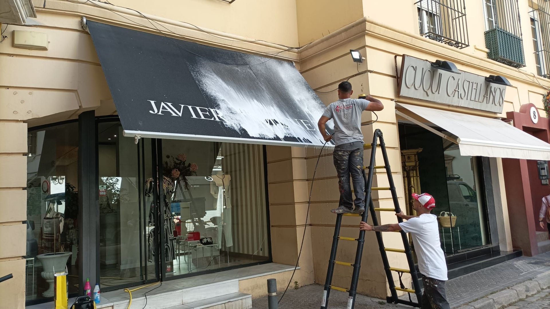 Dos hombres pintando un toldo negro. Uno de ellos, subido a una escalera, aplica pintura blanca. Fachada de una tienda en una calle soleada.