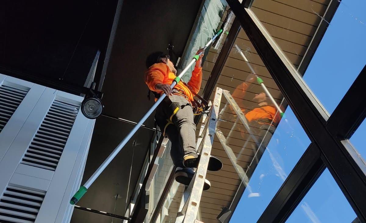 Limpiacristales subido a una escalera, utilizando una rasqueta de mango largo para limpiar una ventana alta. Chaleco naranja.