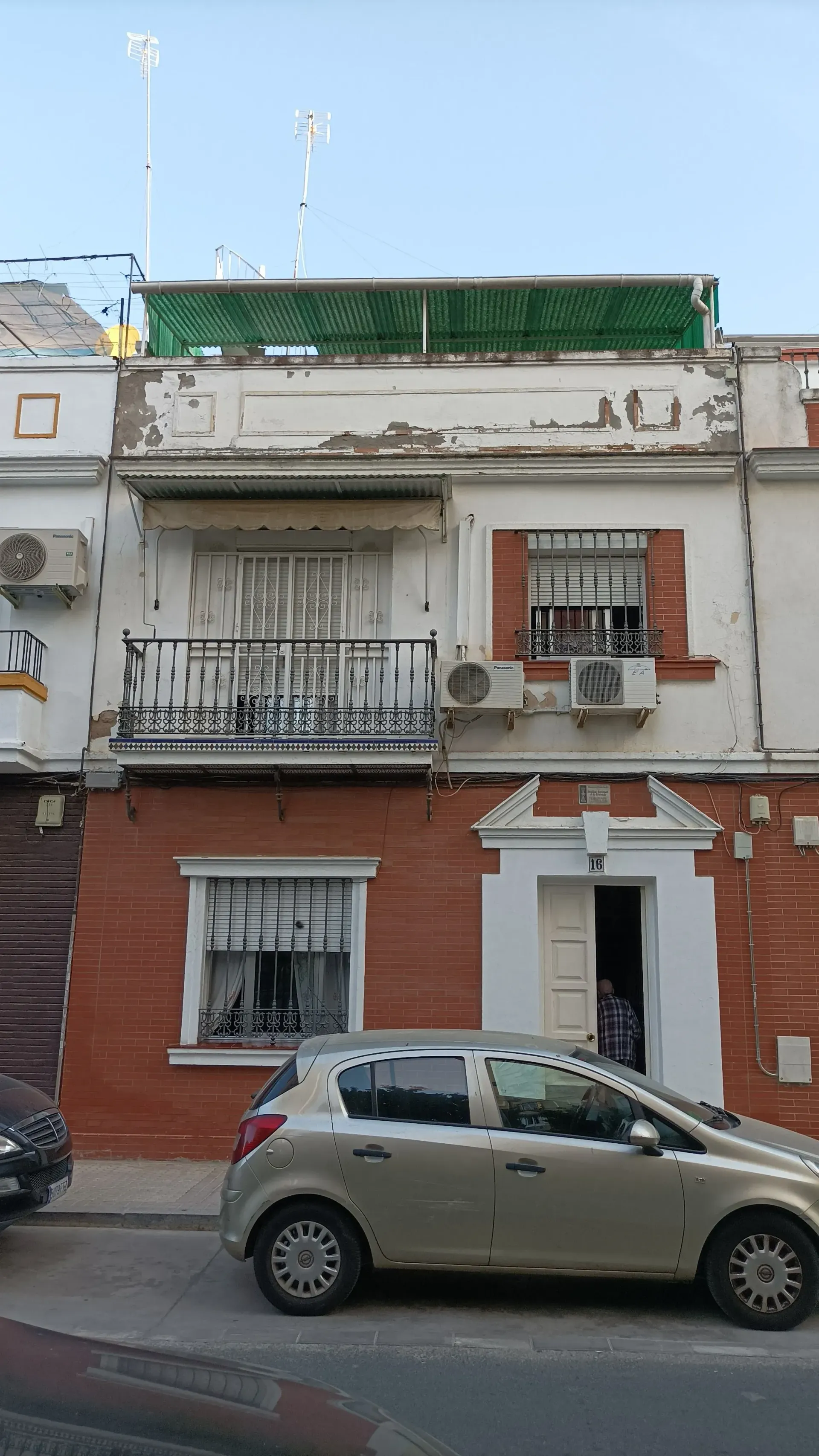 Edificio de ladrillo de dos plantas con balcón, puerta abierta y un coche aparcado delante.