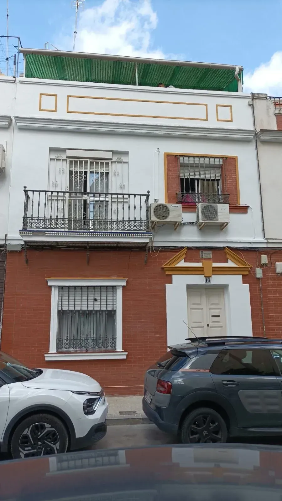 Edificio de dos plantas de ladrillo y blanco con balcón, coches aparcados delante.