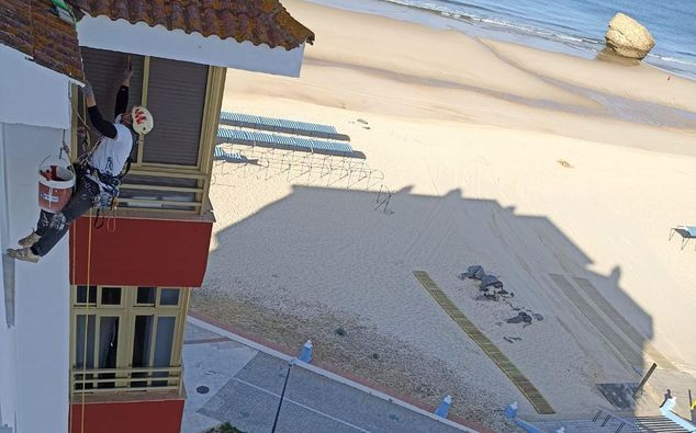 Persona descendiendo en rappel por un edificio, pintando cerca de la playa, con equipo de seguridad y un cubo de pintura.