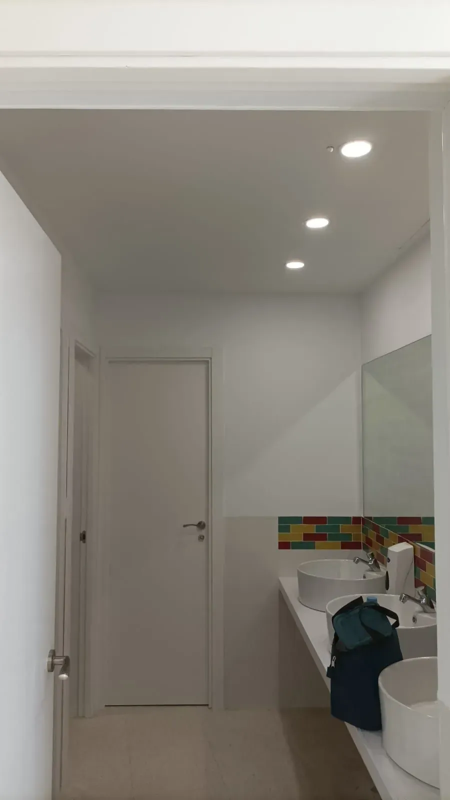 Baño con puerta blanca cerrada, tres luces de techo y lavabo con pared de azulejos de colores.