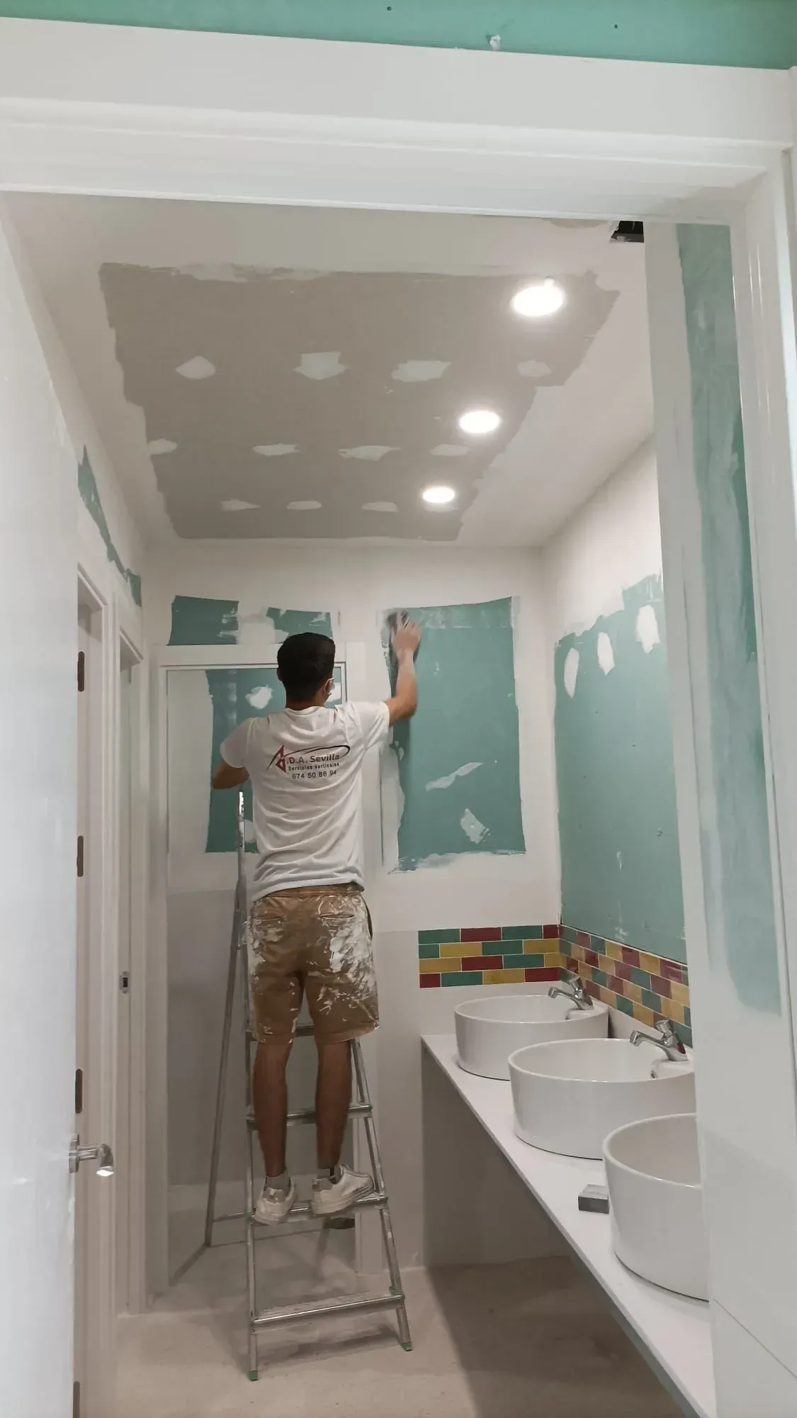 Hombre subido a una escalera enyesando paredes y techo en un baño con luces empotradas; tonos blancos y aguamarina.