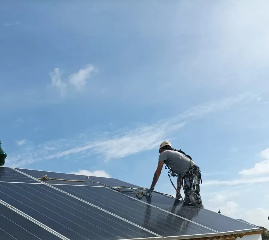 Persona con arnés de seguridad instalando paneles solares en un tejado, bajo un cielo azul brillante.