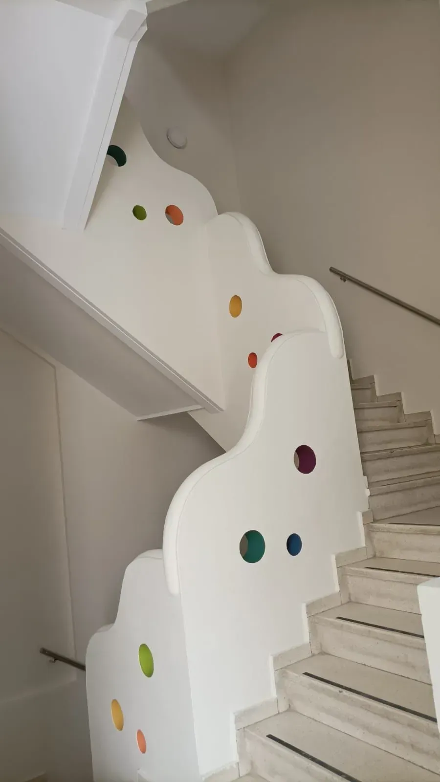 Escalera con pared lateral blanca y ondulada con coloridos recortes circulares. Peldaños de mármol.