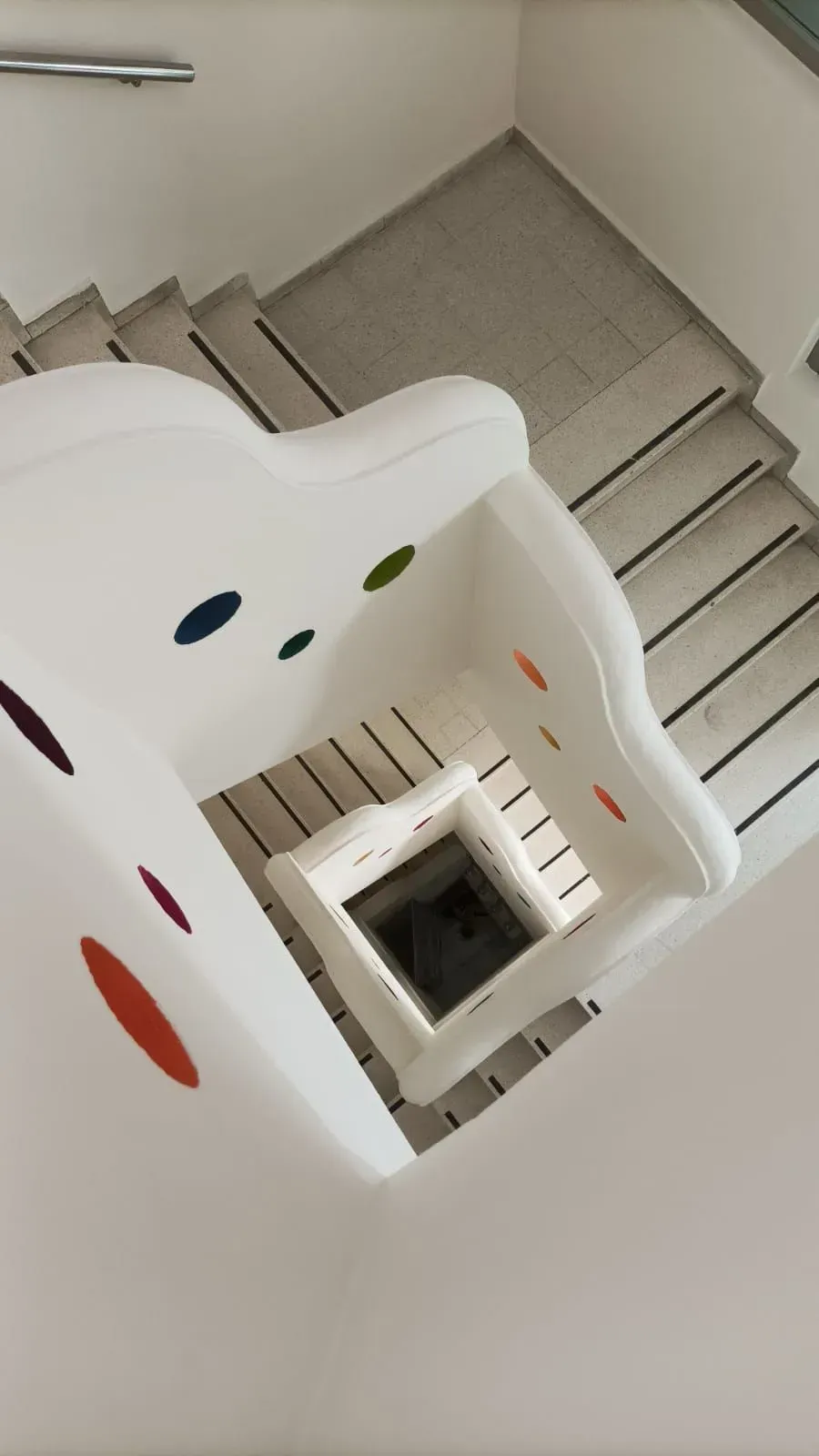 Escalera con barandilla blanca decorada con puntos de colores. Los escalones descienden hasta una abertura cuadrada.