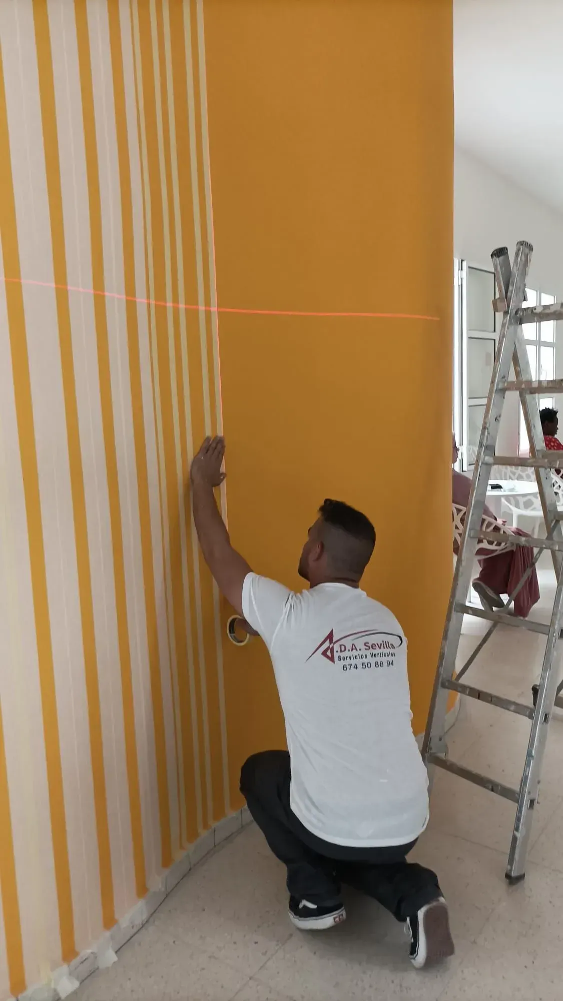 Persona utilizando un nivel sobre una pared con papel pintado a rayas y una sección amarilla lisa.