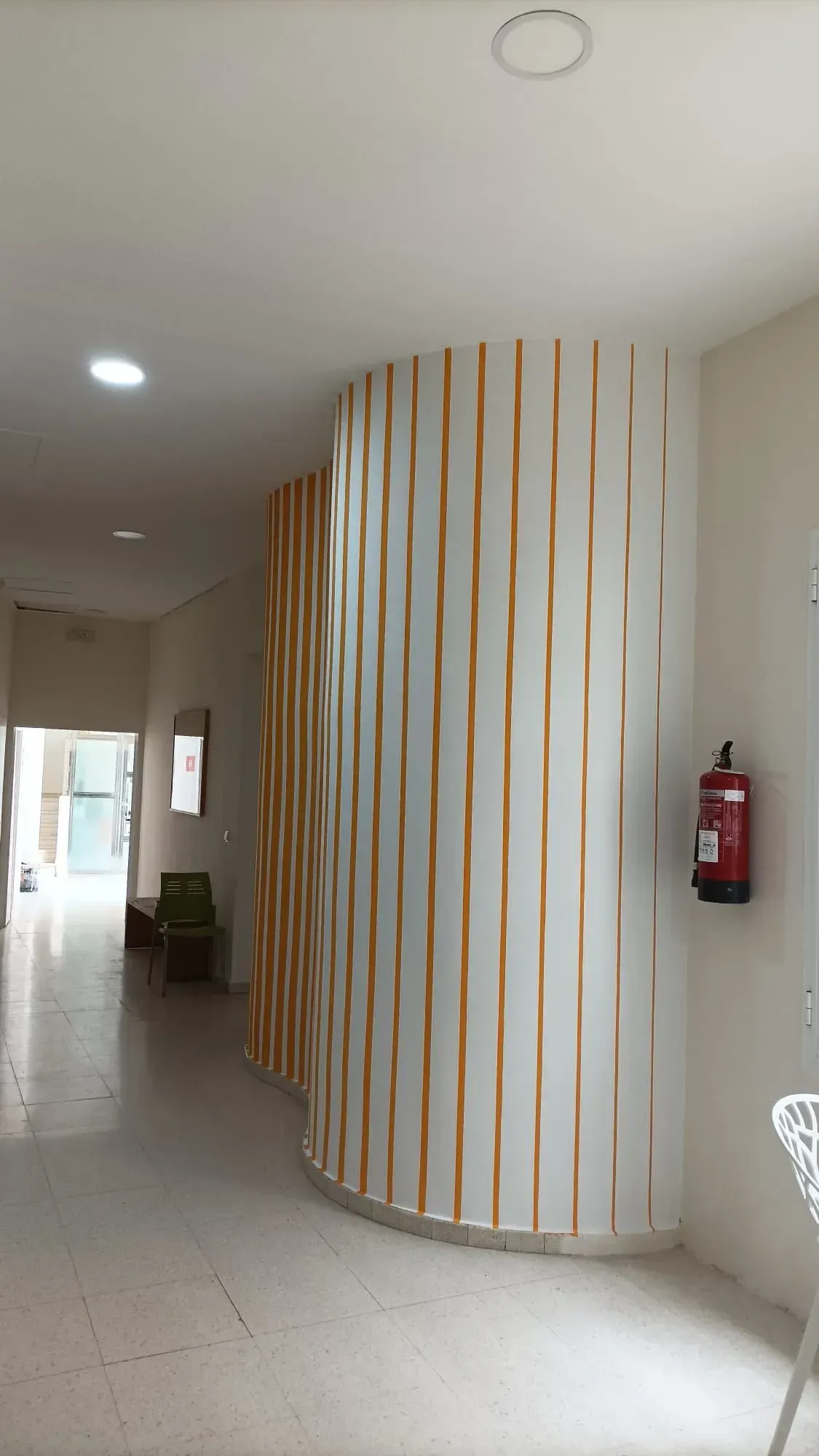 Pasillo con pared blanca curva acentuada por listones verticales de madera; extintor a la derecha.