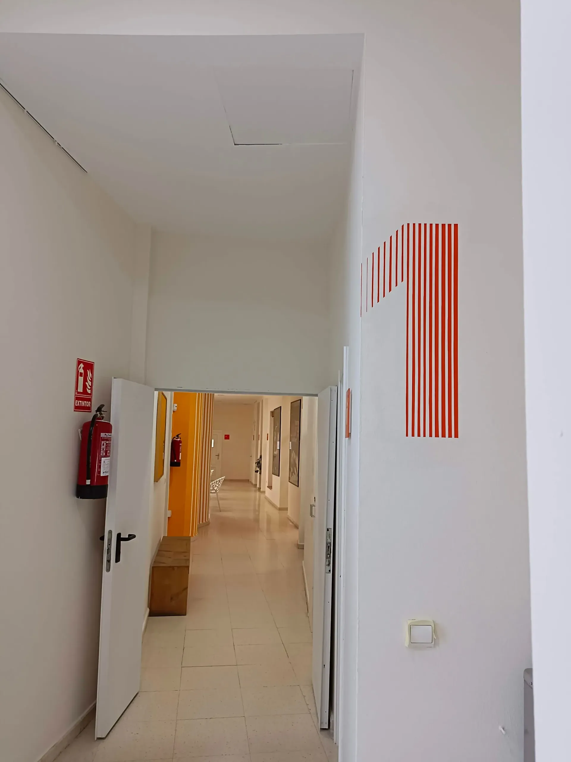 Pasillo con paredes blancas, puertas abiertas, extintor y un gran