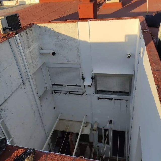 Vista aérea de un patio de paredes blancas con dos ventanas con contraventanas, una estructura metálica debajo y un tejado de tejas rojas.