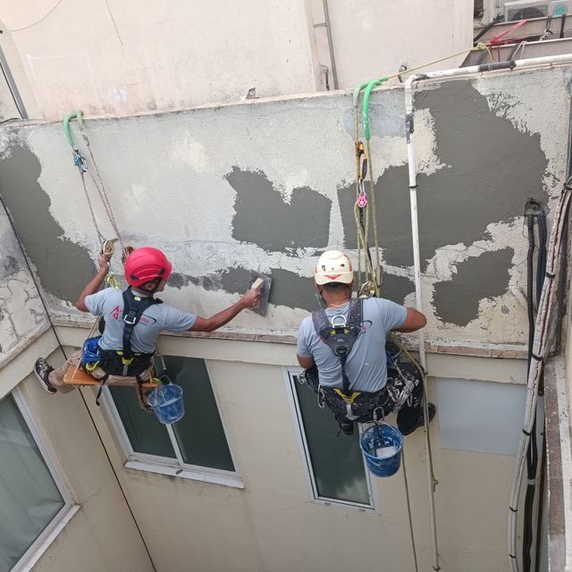 Dos trabajadores, suspendidos de cuerdas, enlucinan el exterior de un edificio. Llevan cascos, arneses y utilizan llanas.