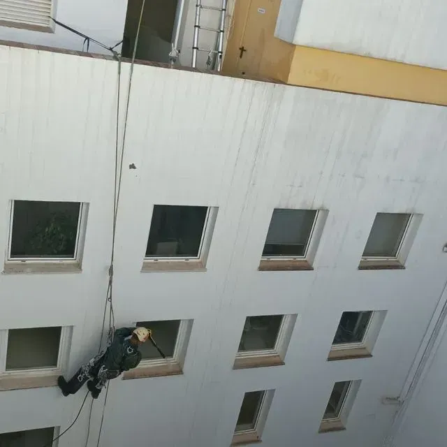 Una persona desciende en rappel por un edificio, vestida con ropa oscura y casco; vista en ángulo desde abajo.