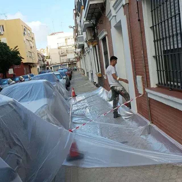Una persona pinta el exterior de un edificio. Un plástico cubre la acera y los coches aparcados en una calle de la ciudad.