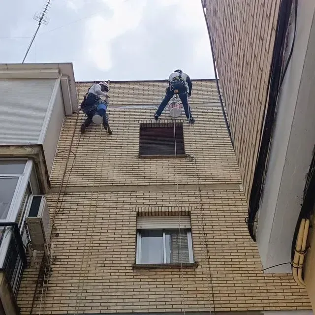 Dos trabajadores sujetos con cuerdas reparan la fachada de un edificio de ladrillo.