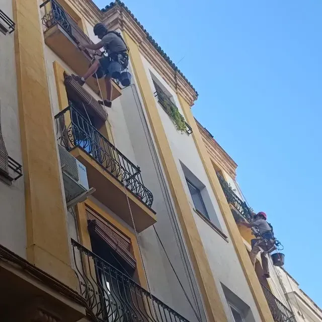 Dos trabajadores colgados de cuerdas, limpiando un edificio amarillo y blanco con balcones contra un cielo azul.