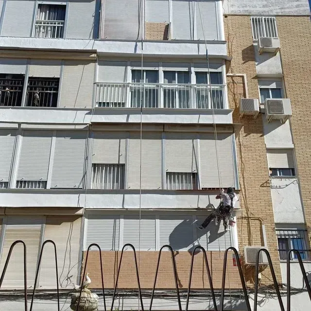 Persona descendiendo en rappel por un edificio, aplicando material en el exterior. Edificio beige con persianas blancas.